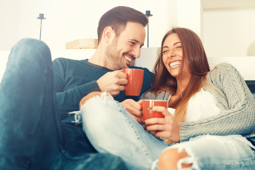 Couple sit close to each other on a sofa having coffee, smiling and laughing, experiencing ease and closeness together.