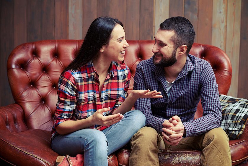 Couple sit on a red leather sofa talking, feeling grounded and confident as partners.