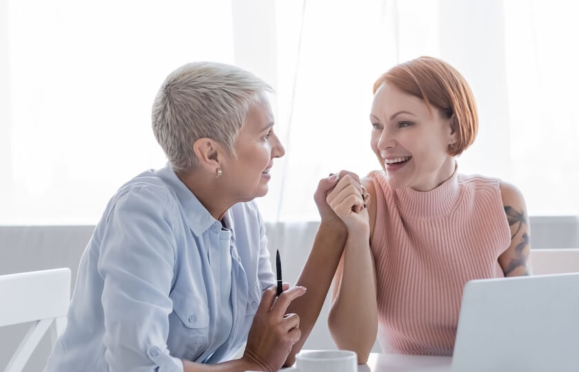 A newly married couple sit at a desk with a laptop, they are facing each other, holdings hands and smiling, representing the relationship dynamics and psychology hub.