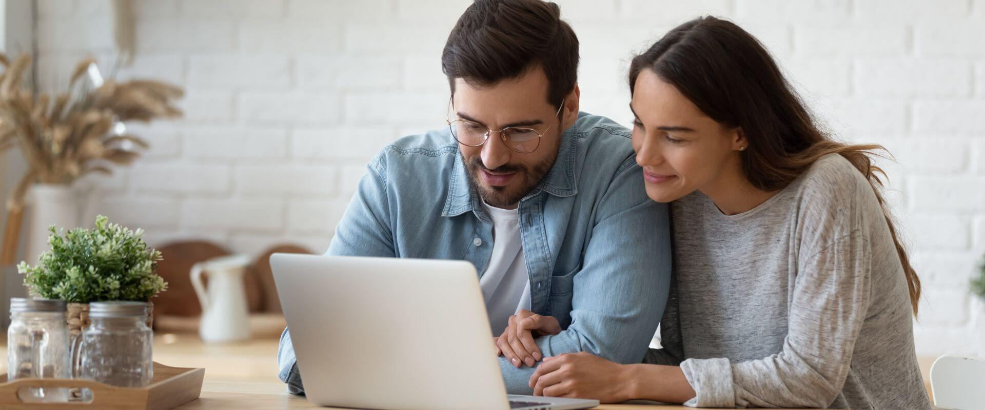 Couple sit a kitchen table reading a blog together on a laptop learning how to create a happy marriage.