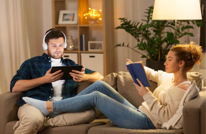 Couple sit on sofa in stress free zone unwinding in their own way reading and listening to music