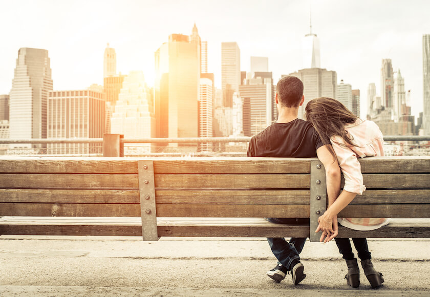 A couple sit on a bench holding hands, watching the sun set on a city scape, reflecting on how conversations feel calmer.