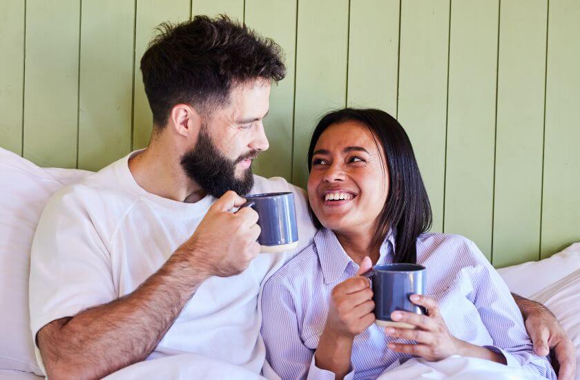 Couple sit in bed drinking coffee as they check in with each other