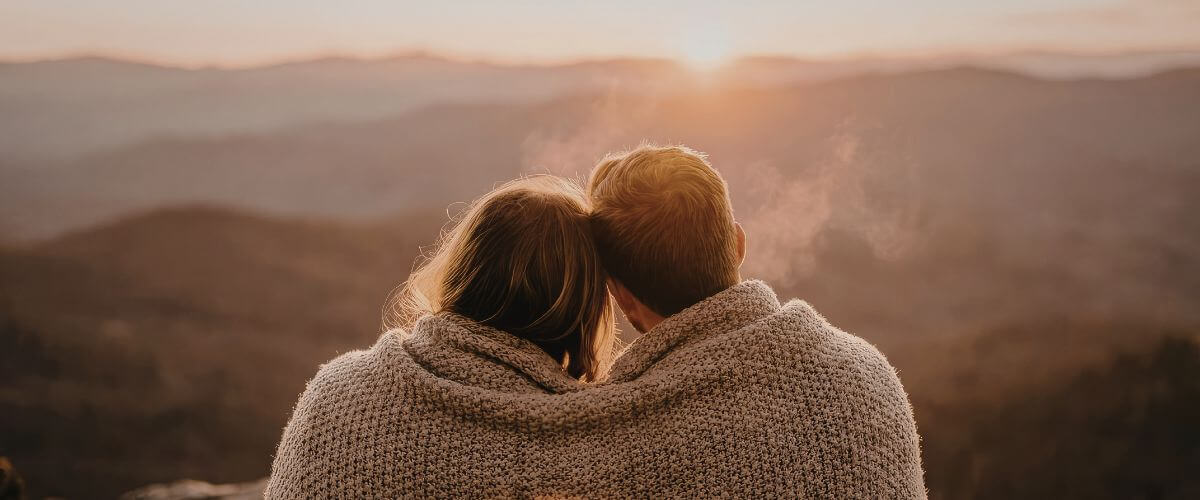 Couple sit with blanket around them on a mountain top looking at the horizon, replying on each other in marriage